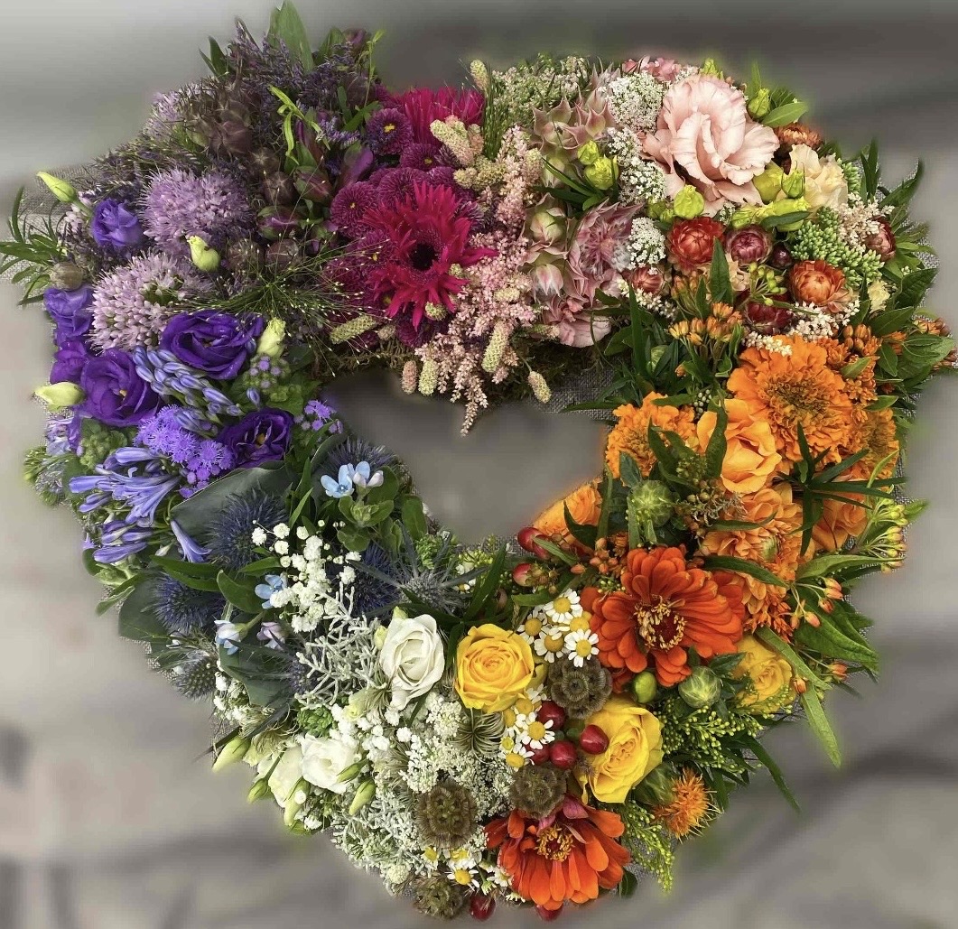 Herzförmiger Trauerkranz mit farblich gruppierten Blüten in Violett, Rosa, Orange, Gelb, Weiß und Blau.