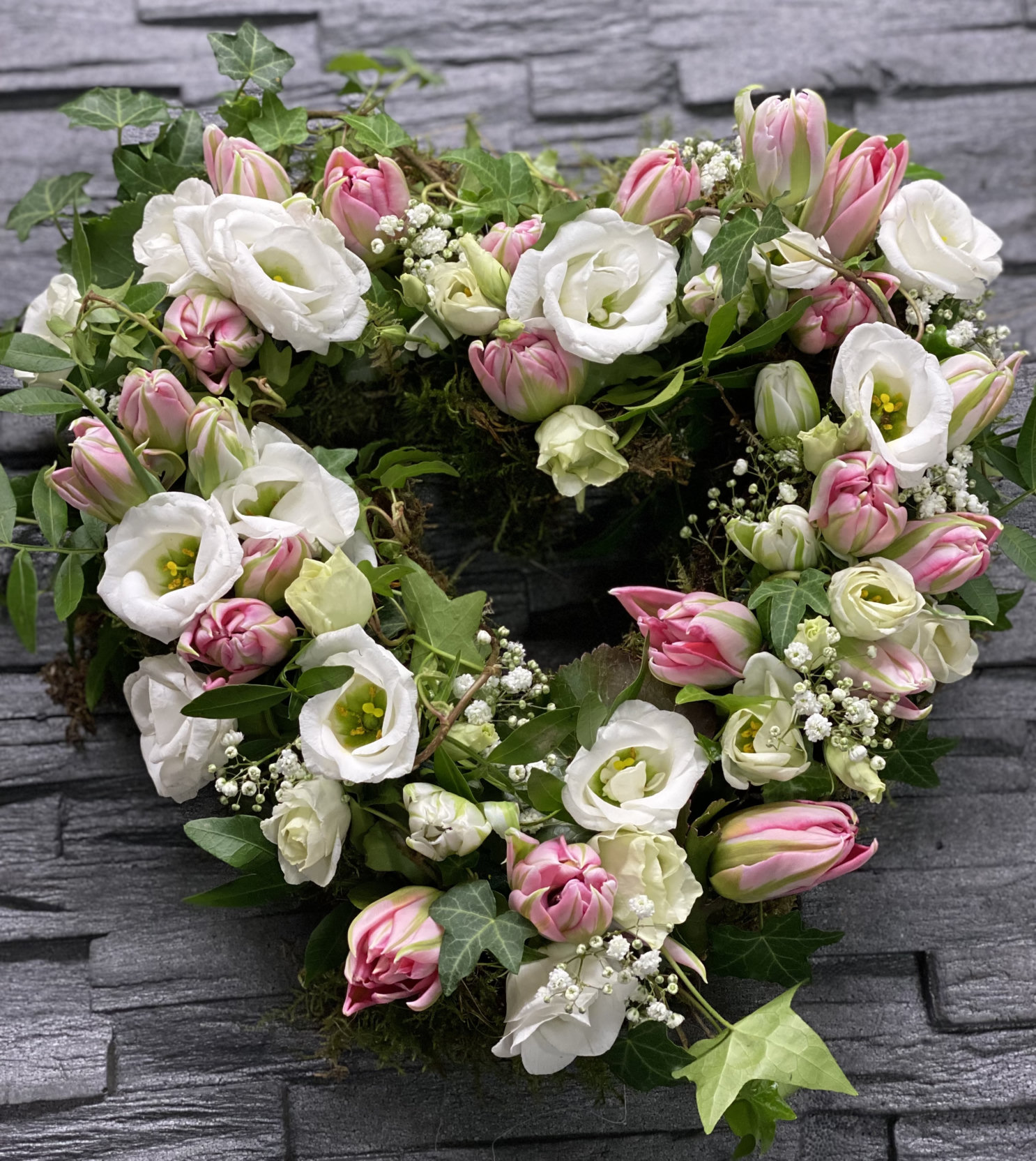 Herzförmiger Mooskranz mit rosa Tulpen, weißen Blüten, Schleierkraut und Efeu vor einer grauen Steinwand.