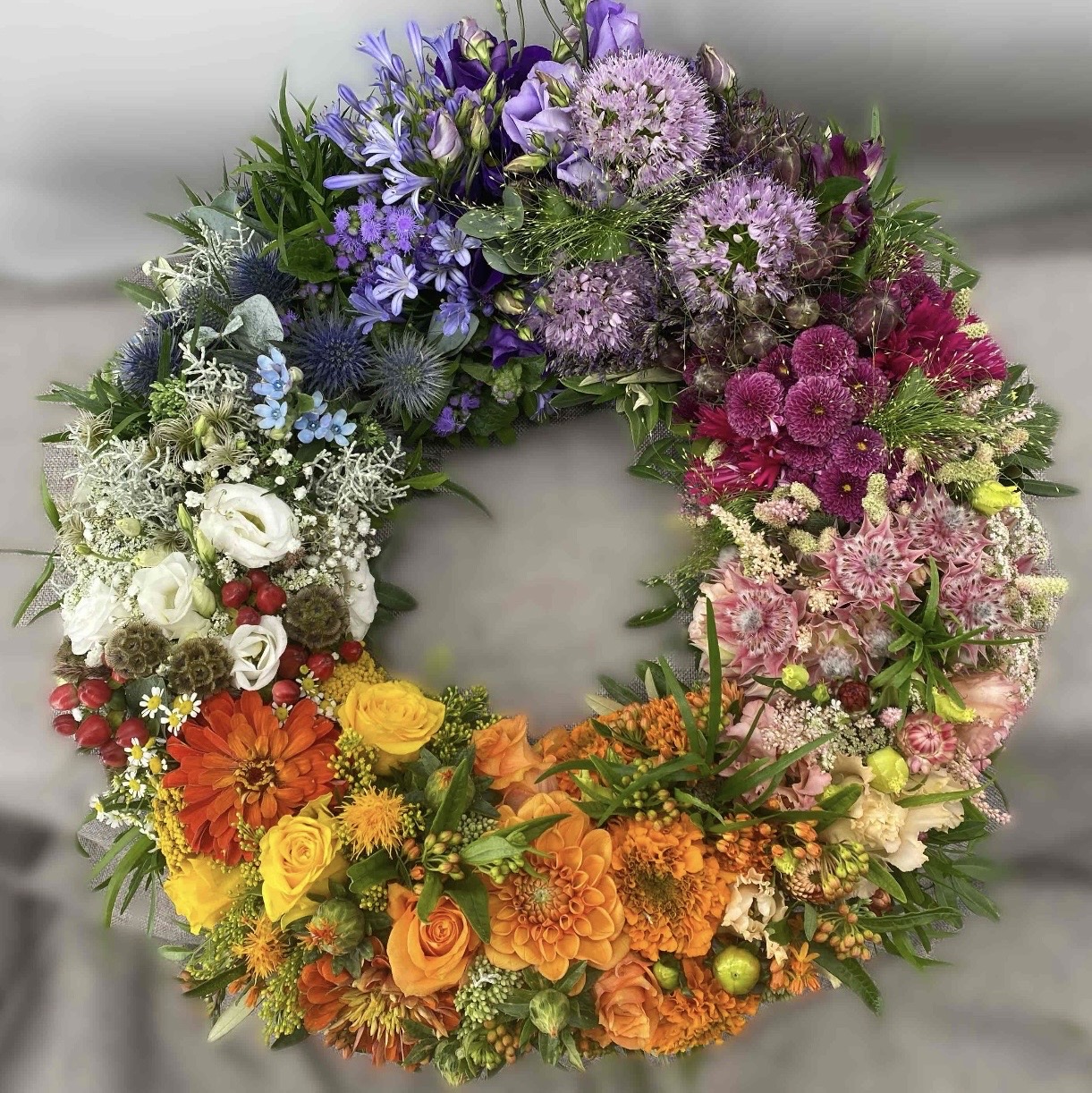Runder Trauerkranz mit dicht gesteckten Blüten im Farbverlauf von Violett über Rosa und Orange bis Gelb und Weiß.